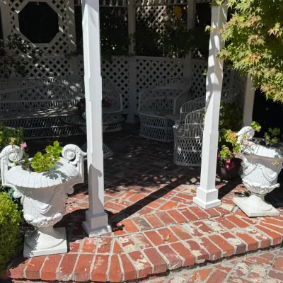 Pair of white cast iron garden urn planters with scroll handles, flanking a brick patio entrance with white wicker patio furniture...