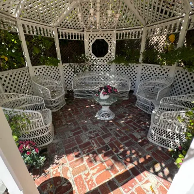 Vintage white wicker patio set including a sofa, four armchairs, and a glass-top coffee table, arranged inside a white wooden gazebo.