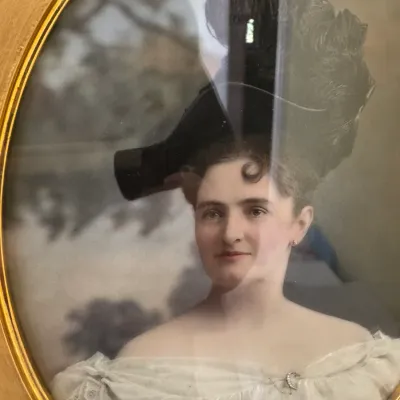 Antique hand-tinted portrait of a Victorian-era woman in an off-the-shoulder lace dress, housed in an oval gold frame with convex glass.