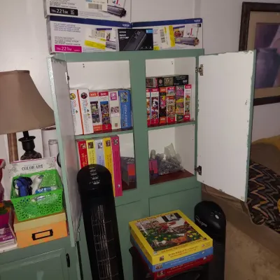 Sage green painted wooden cabinet with open doors, filled with various jigsaw puzzles, flanked by tower fans and other household items.