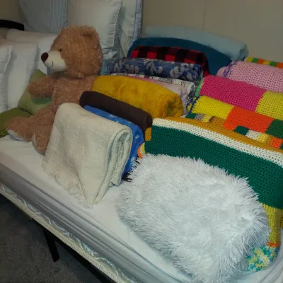 Assortment of colorful blankets, quilts, and pillows arranged on a bed, featuring a large plush teddy bear.