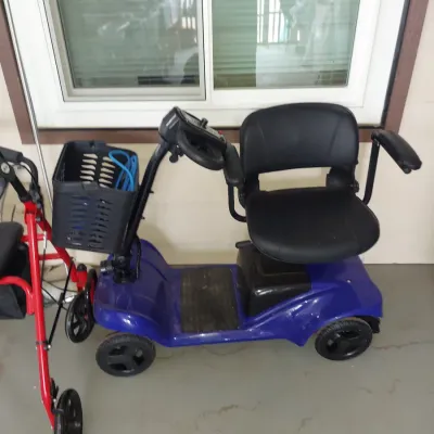 Blue four-wheel mobility scooter with a black seat, armrests, and a front basket, shown alongside a red rolling walker.