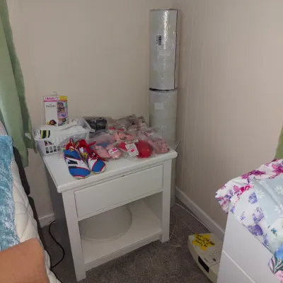 White wooden single-drawer nightstand with an open lower shelf, holding various children's shoes, clothing items, and packaged goods.