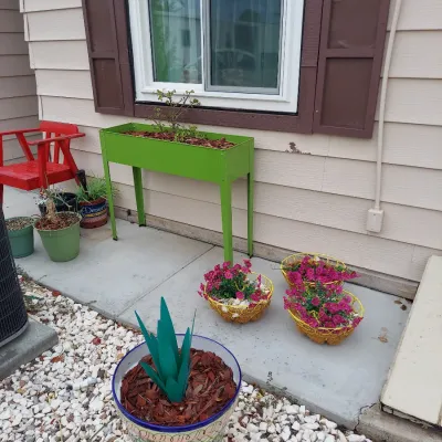 Lime green metal raised garden planter box on legs, shown alongside various potted plants and garden decor on a concrete patio.