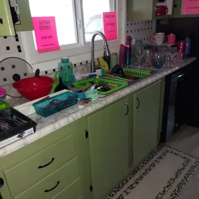 Assorted kitchen utensils, drinkware, and household items displayed for sale on a kitchen counter with green cabinets.