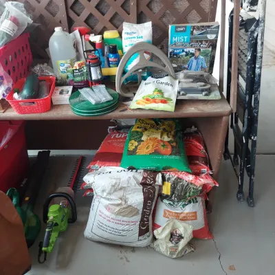 Assorted gardening supplies, including bags of topsoil and garden soil, a hose reel, garden hose, Epsom salt, and a green electric hedge...