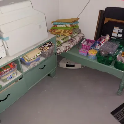 Assorted sewing and crafting supplies, including folded fabrics, storage bins, and a white sewing machine case, arranged on sage green...