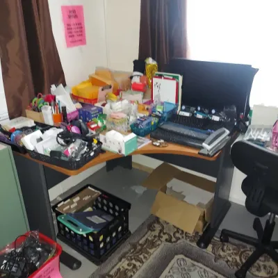 Corner computer desk cluttered with various office supplies, electronics, and storage bins, paired with a black rolling office chair.