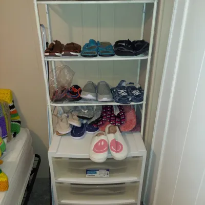 White wire shoe rack with four shelves, positioned over a white plastic three-drawer storage unit, displaying various pairs of shoes,...