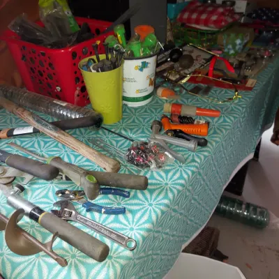 Assorted hand tools and gardening supplies, including pliers, a wrench, hose nozzles, and stakes, displayed on a patterned tablecloth.