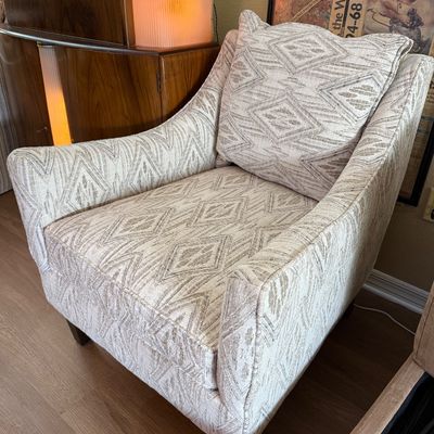 Contemporary upholstered armchair with a neutral geometric diamond pattern, featuring a matching accent pillow and dark wood legs.