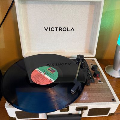 Victrola suitcase-style turntable with a white textured exterior, wood grain faceplate, and a vinyl record on the platter.