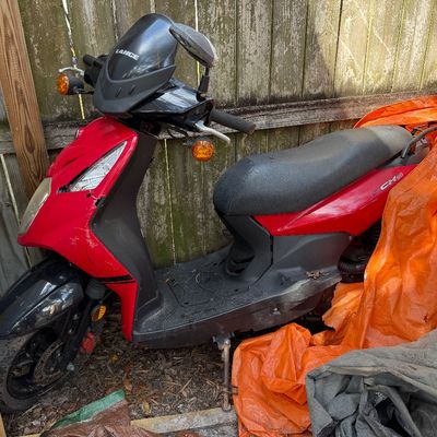 Lance Cabo 50 motor scooter in red and black, showing some wear and damage to the front plastic fairing, parked outdoors next to a wooden...