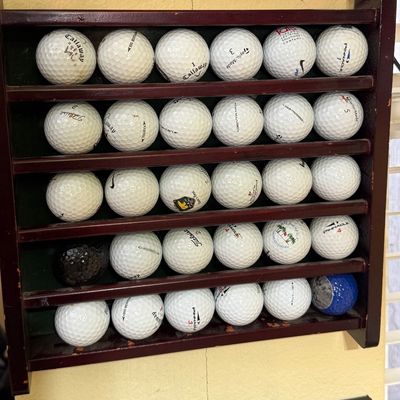 Wooden wall-mounted golf ball display rack holding 28 assorted golf balls, including brands like Callaway, Titleist, and Pinnacle.