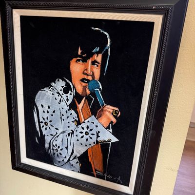 Vintage black velvet painting of Elvis Presley in a white jumpsuit, housed in a dark wooden frame with a fabric liner.