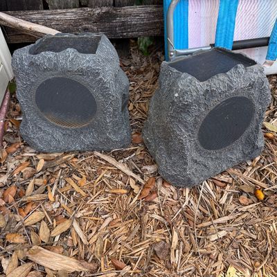 Pair of solar-powered outdoor rock speakers with a textured faux-stone finish, showing some light weathering from outdoor use.