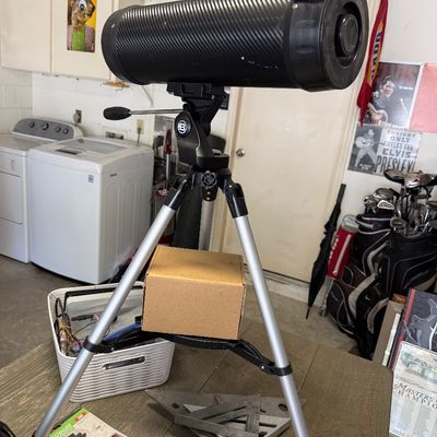 Modern carbon fiber-style telescope mounted on an adjustable silver and black tripod, displayed on a wooden table alongside tools and...