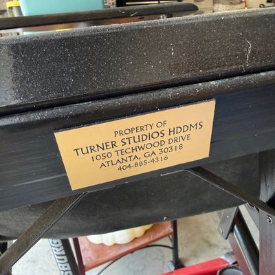A gold-colored property tag reading "Property of Turner Studios HDDMS" affixed to a black metal frame, with a red Pittsburgh...