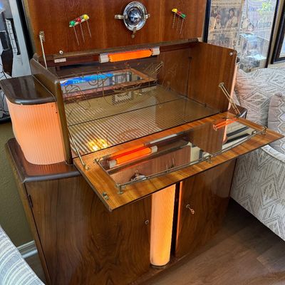 Vintage Art Deco style cocktail bar cabinet featuring a drop-down mirrored mixing surface, integrated orange accent lighting, and a rich...