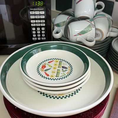 Vintage ceramic pasta bowl set with green trim and vegetable illustrations, shown alongside coordinating mugs and plates.