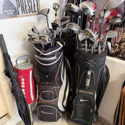 Two golf bags filled with assorted clubs, including a Tommy Armour bag and a Nike bag, accompanied by a red Bag Shag ball retriever and a...