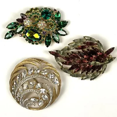 Three vintage costume jewelry brooches, including a green rhinestone floral spray, a multi-toned purple and green leaf-shaped pin, and a...