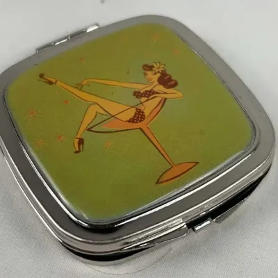 Vintage-style silver-tone compact mirror featuring a retro pin-up girl illustration in a martini glass on a green background.