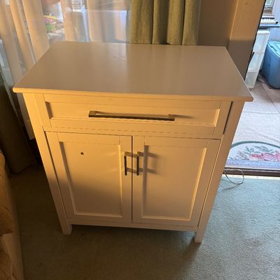 Contemporary white wooden storage cabinet featuring a single top drawer and a two-door lower compartment, accented with modern silver bar...