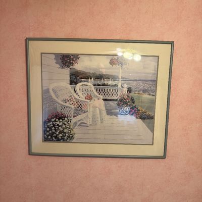 Framed art print depicting a serene porch scene with white wicker chairs overlooking a body of water, presented in a simple grey frame...