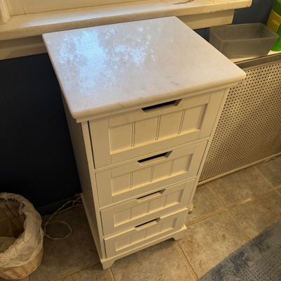 Contemporary white four-drawer storage cabinet featuring a polished marble top and beadboard-style drawer fronts with integrated cutout...