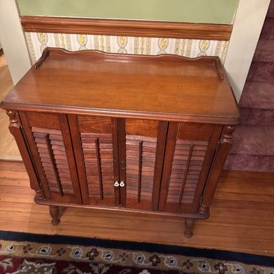 Vintage wooden cabinet featuring louvered doors, turned legs, and a gallery-style top edge, showing some light surface wear.