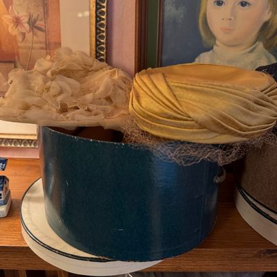 Vintage mid-century women's hats, including a ruffled cream floral hat and a draped gold satin turban-style hat with netting,...