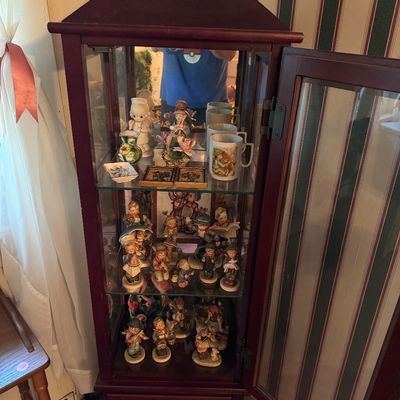 Cherry-finished wooden curio cabinet with a peaked top, glass shelves, and a mirrored back, filled with various porcelain figurines and...