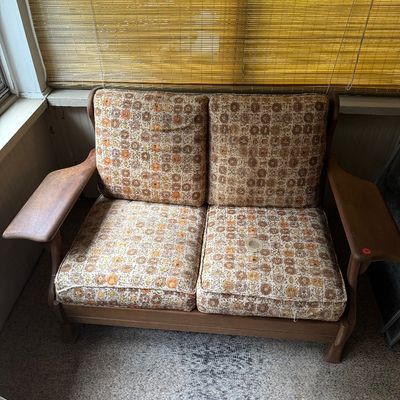 Mid-century modern wooden loveseat with wide paddle arms and patterned earth-tone cushions showing some wear.