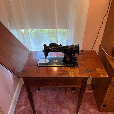 Vintage black Singer sewing machine housed in a wooden cabinet with folding extension leaves, showing some wear to the wood finish.