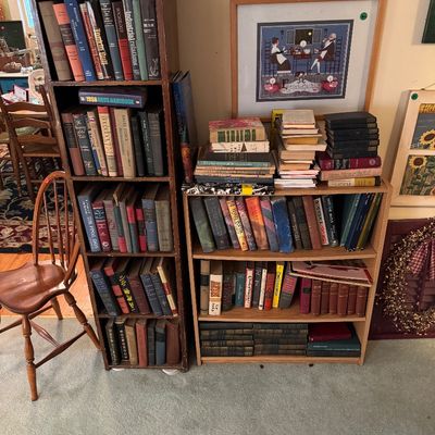 Two wooden bookshelves filled with vintage books, including a tall, narrow dark wood shelf and a shorter, wider light oak shelf,...