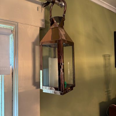 Contemporary copper-finished metal and glass candle lantern, hanging from a curtain rod and containing a white pillar candle.
