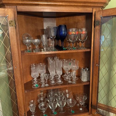 Wooden corner hutch with glass-paneled doors, filled with an assortment of vintage glassware including wine glasses, goblets, and...