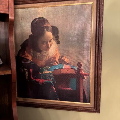 Framed canvas print of Johannes Vermeer's "The Lacemaker," presented in a textured brown wooden frame with a gold-toned...