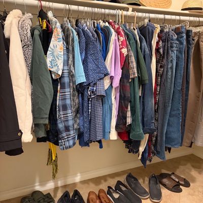 Sale Photo Thumbnail #67: Assorted men's clothing including shirts, jackets, and jeans hanging on a closet rod, with various shoes, sandals, and hats arranged on the shelf and floor.