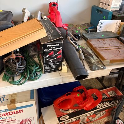 Sale Photo Thumbnail #77: Assorted garage tools and household items, including a Panasonic car vacuum cleaner in its box, a Craftsman leaf blower, a Black & Decker hedge trimmer, and extension cords.