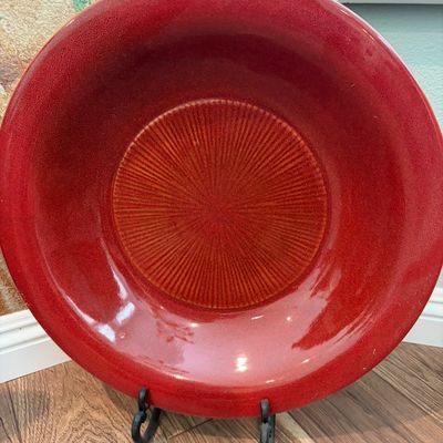 Sale Photo Thumbnail #50: Large ceramic decorative bowl with a vibrant red glaze and a textured, sunburst-patterned center, displayed on a black metal stand.