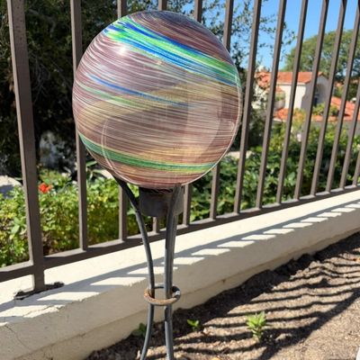 Sale Photo Thumbnail #23: Swirling multi-colored glass gazing ball in shades of brown, green, and blue, displayed on a black metal stand in a garden setting.