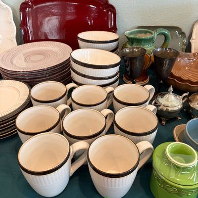 Sale Photo Thumbnail #63: Assorted ceramic tableware collection, featuring a set of white mugs and bowls with dark rims, stacked plates, a red serving platter, and various decorative pottery pieces.