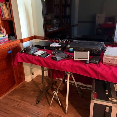 Sale Photo Thumbnail #87: Assorted electronics including a flat-screen television, VCR, DVD player, and portable screens displayed on a red-clothed table alongside tripods and a road case.