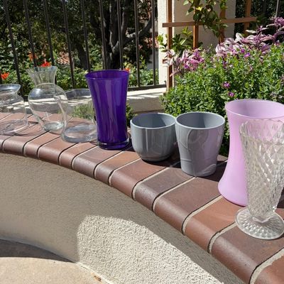Sale Photo Thumbnail #15: Assortment of glass and ceramic vases, including clear glass, vibrant purple, pastel pink, and grey ceramic planters, displayed outdoors on a brick ledge.
