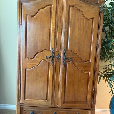 Sale Photo Thumbnail #70: French Provincial style wooden armoire featuring arched double doors with ornate brass hardware, a single bottom drawer, and a carved floral medallion on the top crown.