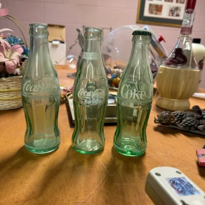 Three vintage green glass Coca-Cola bottles, including embossed and painted label designs, displayed on a wooden table.