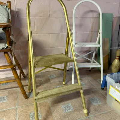 Vintage gold-toned metal step stool with a high arched handle, shown alongside a white metal step stool and a wooden high chair.