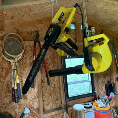 Eager Beaver 14-inch electric chainsaw and a yellow electric leaf blower hanging in a shed alongside vintage tennis rackets and garden...